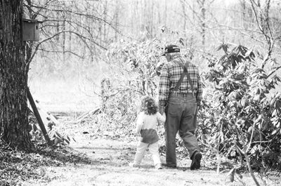 A man in overalls holds a younger girl's hand while walking in a backyard. This represents the beloved collection of greeting cards by beknown.