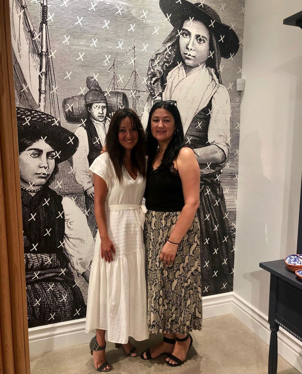 Nancy Fernandes and Fatima Santos, Founders of Saudade Toronto, standing in front of full length mural wallpaper of female artisans. Nancy is the inspiration for the greeting card Saudade, part of the bel loved collection by beknown.